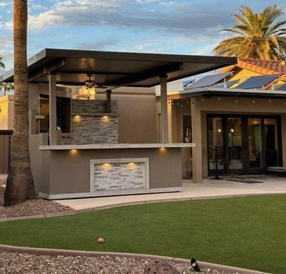 Outdoor kitchen bar unit installed on a patio in a modern single-story house, with a covered seating area, stone tile backsplash, and artificial grass lawn in the foreground.