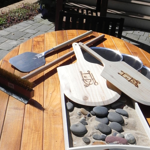 Wooden pizza peel, brush, and stone tray on a wooden table with pebbles.