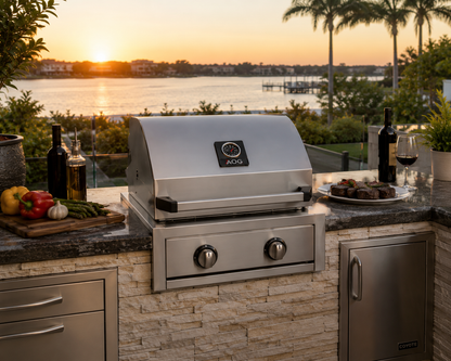 American Outdoor Grill AOG 24-inch T-Series built-in propane grill installed in a luxury outdoor kitchen with stone island, stainless steel cabinetry, and waterfront sunset view