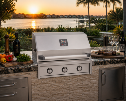 American Outdoor Grill 30-inch T-Series built-in stainless steel gas grill installed in a luxury outdoor kitchen with granite countertops, stone island, waterfront sunset view, premium outdoor cabinetry, and gourmet entertaining setup.