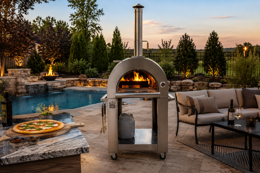 Luxury outdoor patio featuring a Kokomo stainless steel pizza oven with live wood-fired flame beside a resort-style pool, stone fire feature, and evening entertaining lounge area.