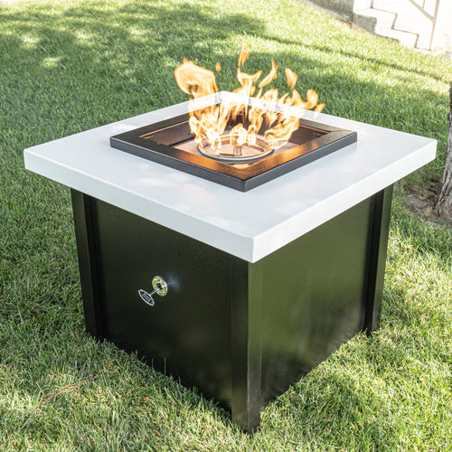 Kamoa square metal fire table with white top and black base, featuring circular burner with active flames for luxury outdoor entertaining