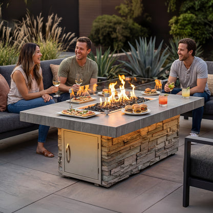 Luxury outdoor entertaining space featuring the Cal Flame Mojave RT601M rectangular gas fire pit table with cultured stone base, porcelain tile top, glass wind guard, black lava rock fire media, and modern patio seating with guests gathered around the flames at dusk.