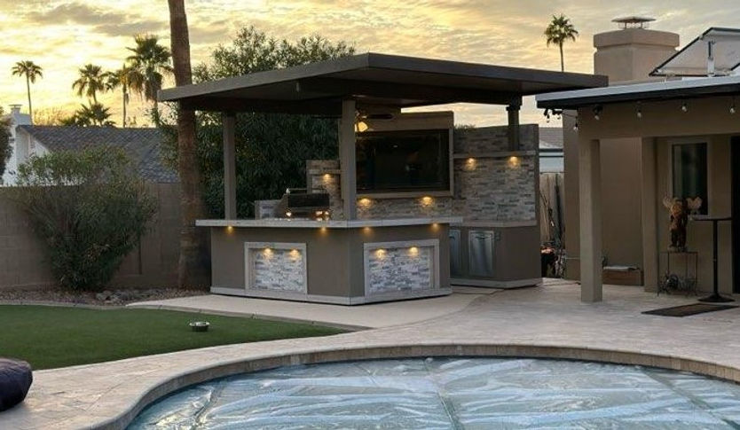 Outdoor kitchen with TV and patio lights near a pool at sunset.